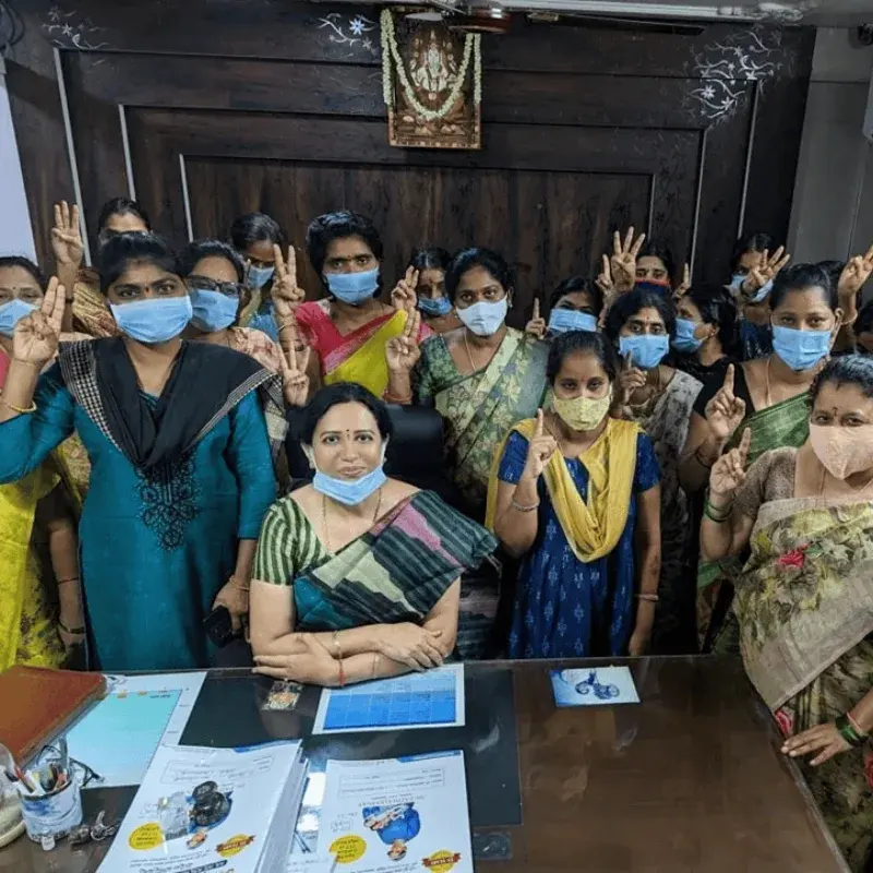 Dr. Padmaja seated at her desk with a large group of patients standing behind her, many raising their hands in celebration